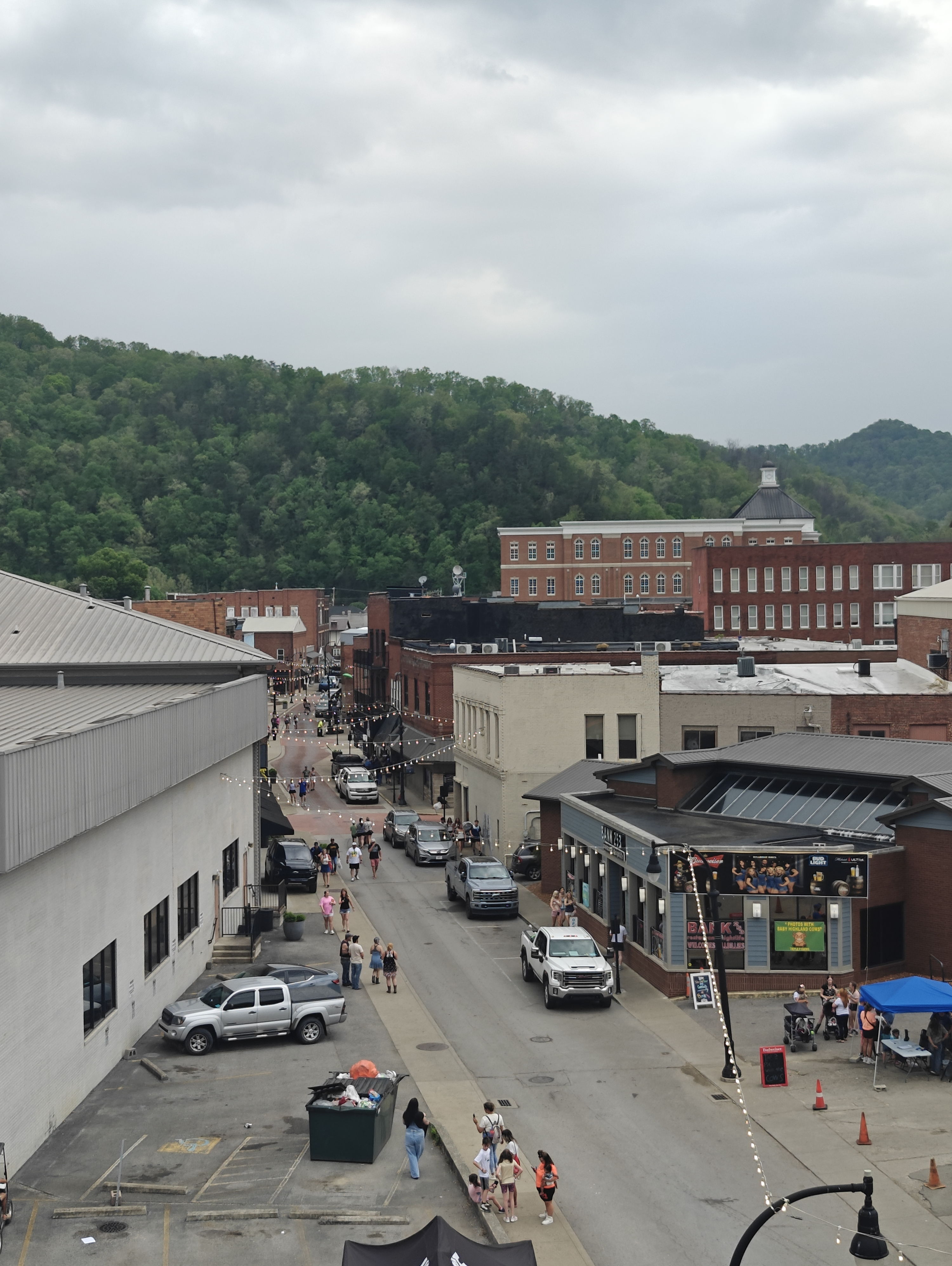 A quaint town street with parked cars, people, and overcast skies, evoking a calm, community atmosphere amid rolling hills.