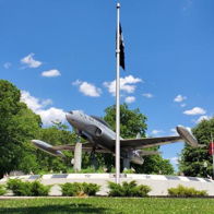 Military aircraft on a pedestal, surrounded by flags, set against a blue sky, symbolizing patriotism and honor.