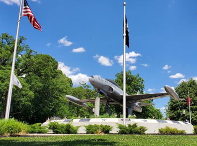 Military aircraft on a pedestal, surrounded by flags, set against a blue sky, symbolizing patriotism and honor.