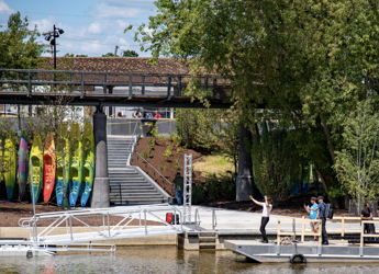 Colorful kayaks line a dock by a serene water body, where people enjoy a sunny day in a vibrant, green setting.