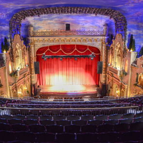 Grand theater interior with ornate decor, a vibrant stage, and deep red curtains, evoking anticipation for a live performance.