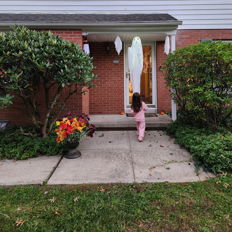 A charming suburban home with a child in pink, festive decorations, green shutters, and colorful flowers evokes joy and warmth.