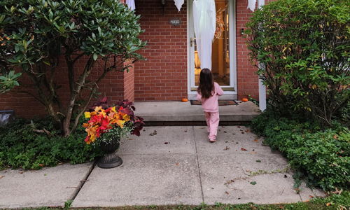 A charming suburban home with a child in pink, festive decorations, green shutters, and colorful flowers evokes joy and warmth.