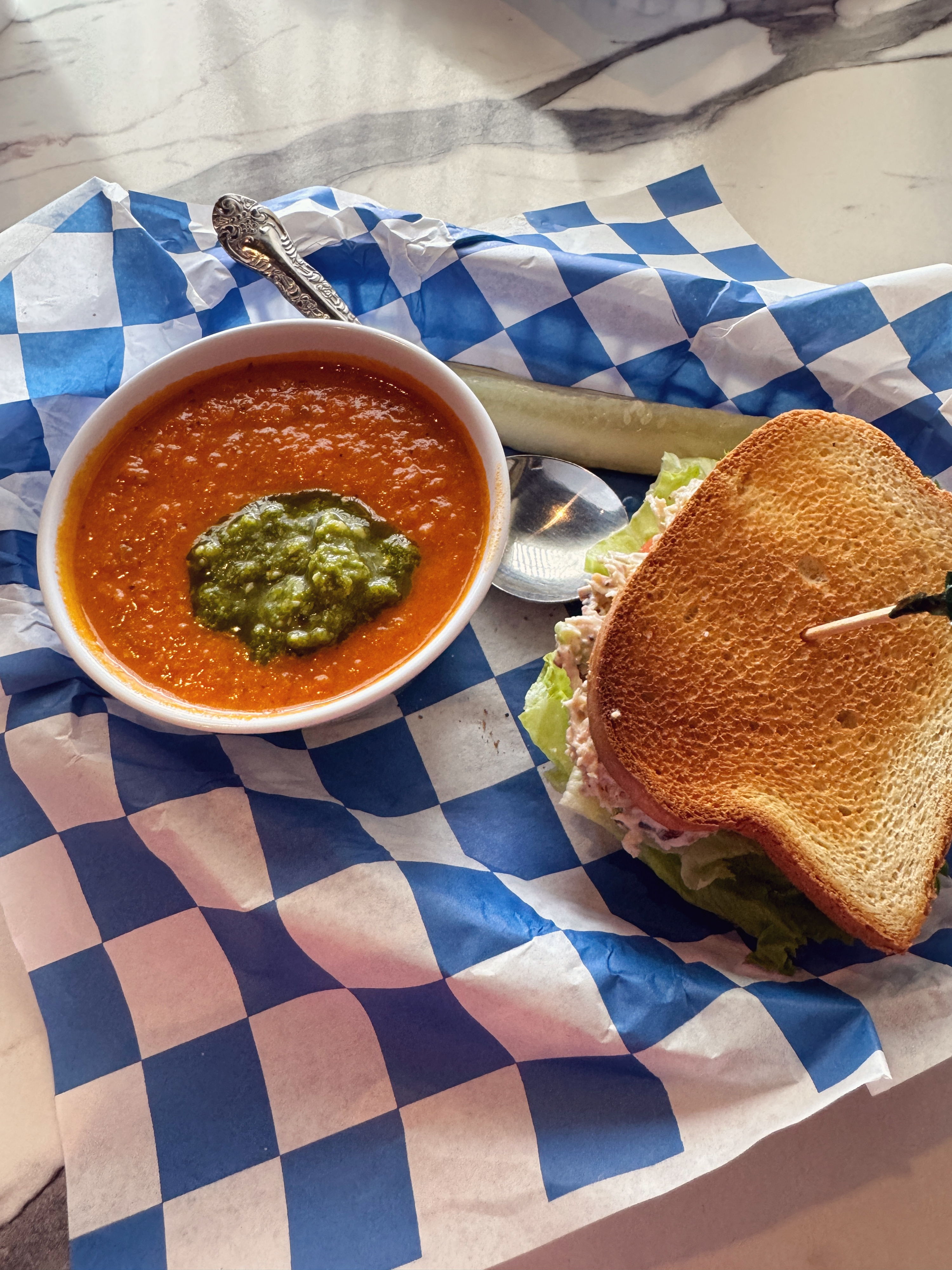 Bowl of tomato soup with pesto, accompanied by a toasted sandwich, served on checkered paper for a cozy, inviting lunch.