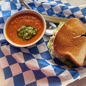 Bowl of tomato soup with pesto, accompanied by a toasted sandwich, served on checkered paper for a cozy, inviting lunch.