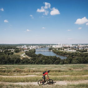 Mountain biker on a hillside path with a river and town below, under a bright sky, capturing adventure and nature's beauty.
