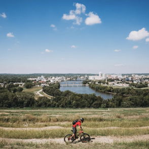 Mountain biker on a hillside path with a river and town below, under a bright sky, capturing adventure and nature's beauty.